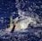 A large fish is caught on a fishing line, thrashing around in deep blue water, creating splashes and bubbles. The fish has a shiny, iridescent body with prominent fins and is attached to a lure with an orange tip.