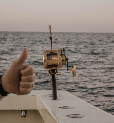 A boat fishing trip on the open sea with a fishing reel mounted on the side of the boat. The ocean stretches to the horizon under a clear sky, and a person gives a thumbs-up gesture, indicating approval or success.