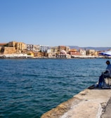 A person is sitting under a blue and white umbrella, fishing along a waterfront promenade. The background consists of a picturesque town with colorful historic buildings and a clear blue sky. Bicycles and street lamps line the path.