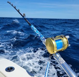 A fishing rod with a gold reel is mounted on the side of a boat, extending over the deep blue ocean. The waves create white foam as the boat moves forward under a clear blue sky.