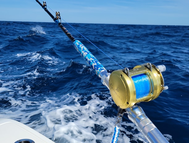 A fishing rod with a gold reel is mounted on the side of a boat, extending over the deep blue ocean. The waves create white foam as the boat moves forward under a clear blue sky.
